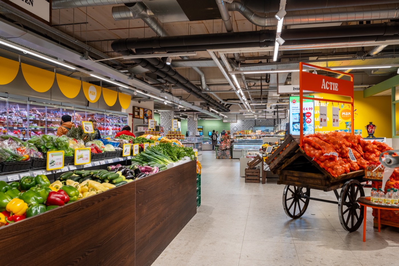 De groente- en fruitafdeling met paprika's en courgettes in houten bakken, en een oude houten kar vol netten uien onder het oranje actiebord.