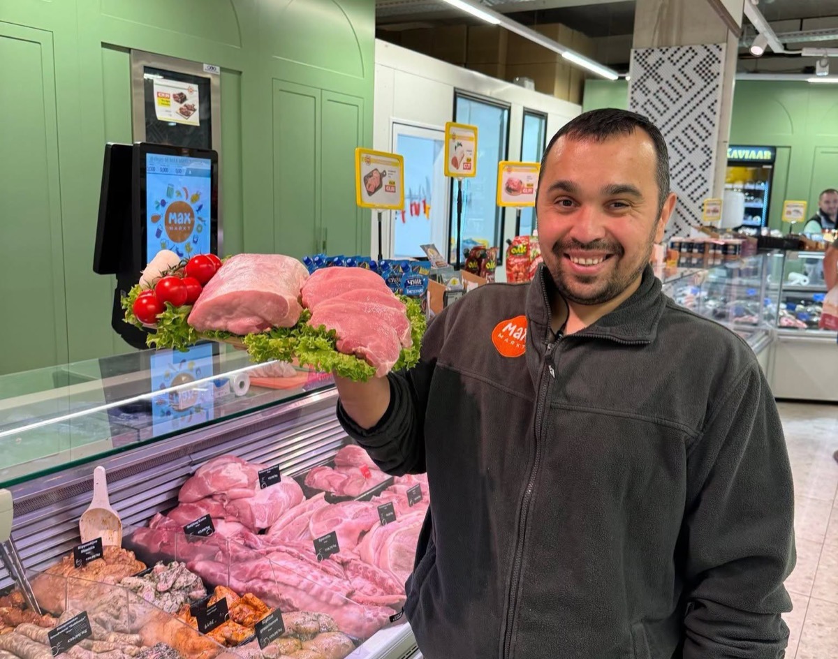 Onze slager houdt lachend een schaal met verse varkenslapjes omhoog naast de vitrine van de slagerij.