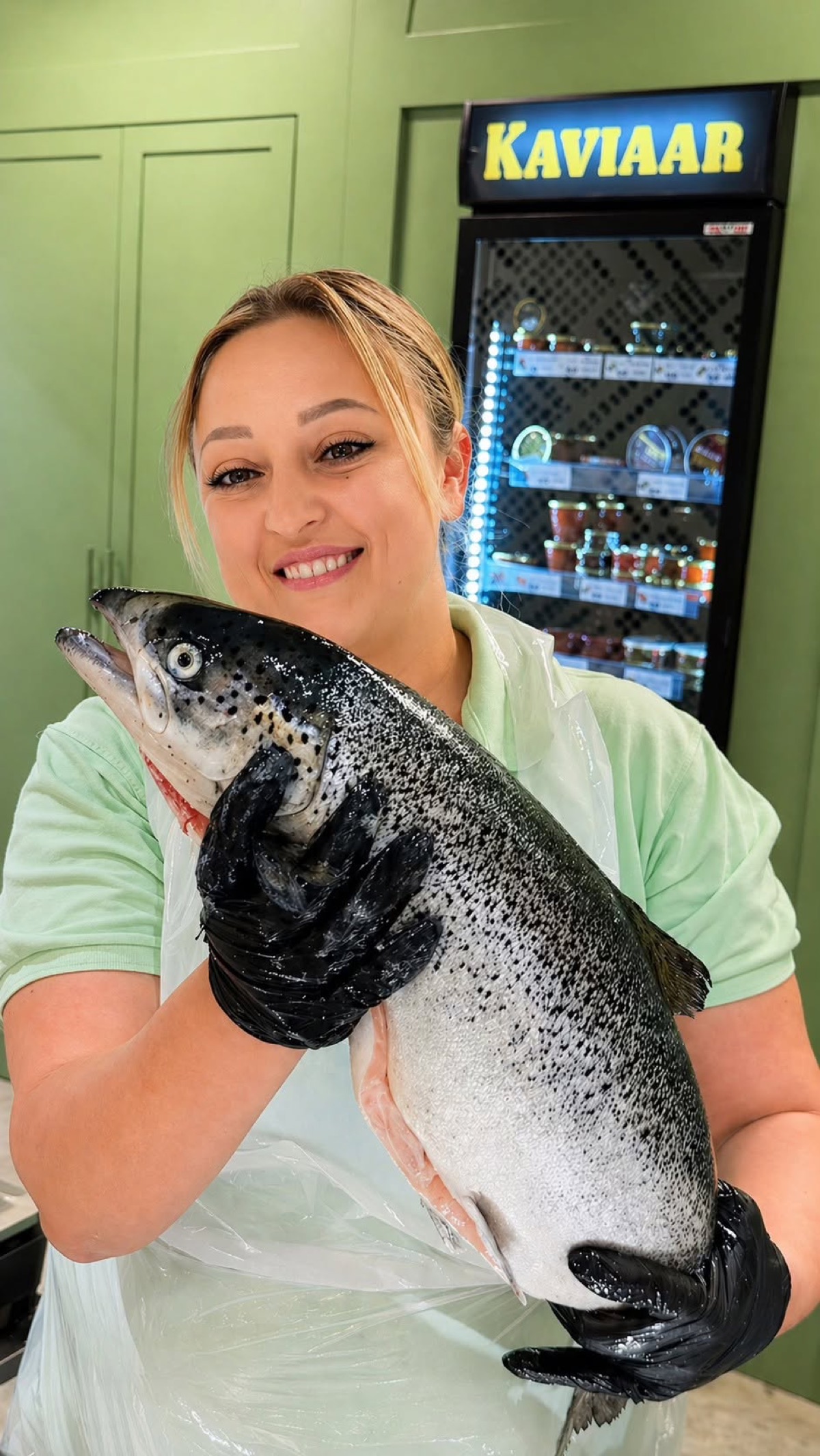 Een collega van de visafdeling houdt lachend een hele verse zalm vast, voor de verlichte kaviaarkoelkast en de groene kasten van de toonbank.