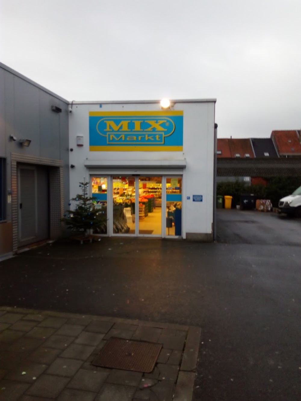 De winkel in Gent op een grijze winterochtend: het blauwgele Mix Markt-bord boven de schuifdeuren, binnen de verlichte schappen en een kerstboom naast de ingang.