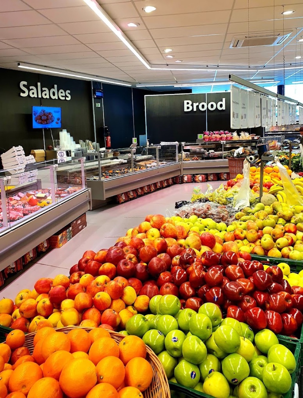 Binnen in Mortsel: bergen appels, sinaasappels en peren in de groente- en fruitafdeling, met achterin de toonbanken onder de borden Salades en Brood.